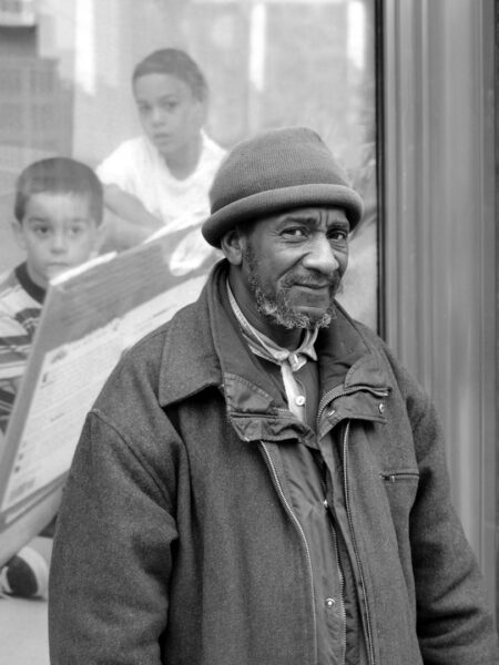 Black man wearing a wool cap and winter coat standing in front of a window with two children in the background watching the scene.