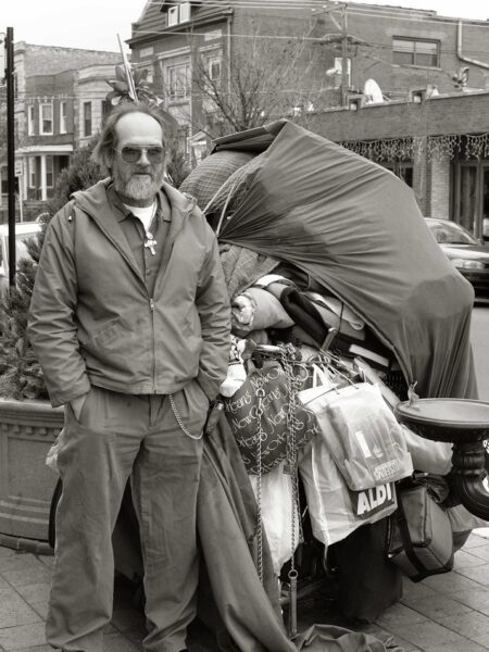 Man in sunglasses standing next to a shopping cart overloaded with bags, blankets, and his life's possessions on the street.