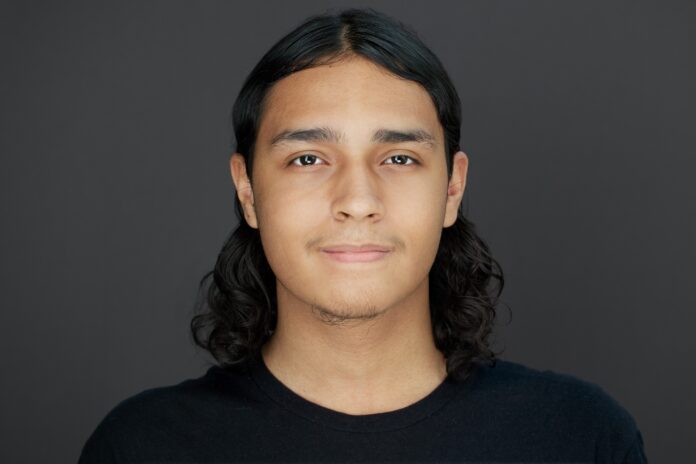 Headshot of young man with long black hair against a dark backdrop.