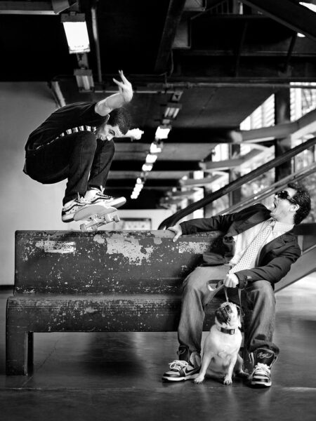 A black and white photo capturing a skateboarder mid-jump over a bench where a seated man wearing sunglasses is expressionlessly looking up at the skateboarder, with a small dog on a leash sitting between the man's legs.