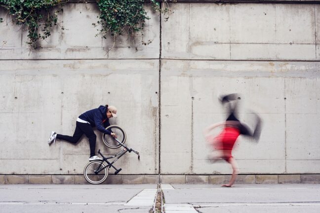 A person appears frozen in an unusual pose while fixing a bicycle against a concrete wall, with another person running past in a blur, showcasing movement.