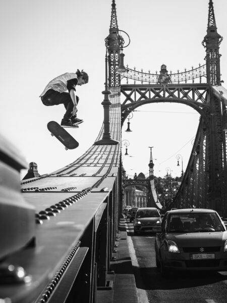 A skateboarder performs a trick in the air above a ramp installed on a bridge, with ornate street lamps and cars driving on the roadway below, captured in black and white.