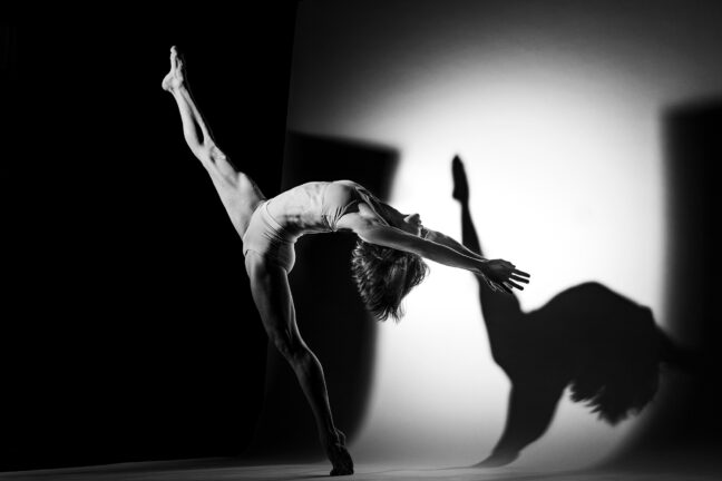 Black and white image of a female dancer performing a backbend with one leg raised high, casting a shadow on the wall behind her.