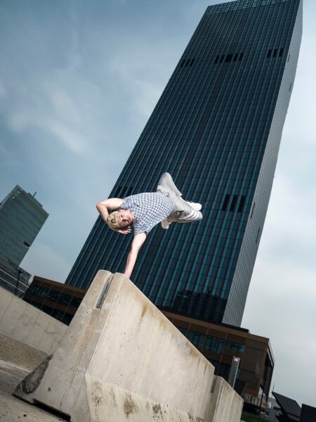 A man performs a one-hand handstand on a concrete block with a tall glass building in the background, creating a dramatic urban scene with a clear sky.