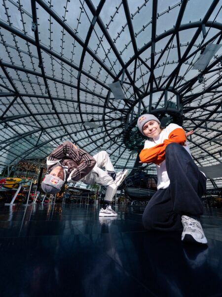 Two people in a modern glass-roofed building, with one person balancing upside down on their head and another person squatting with arms crossed, both smiling at the camera.