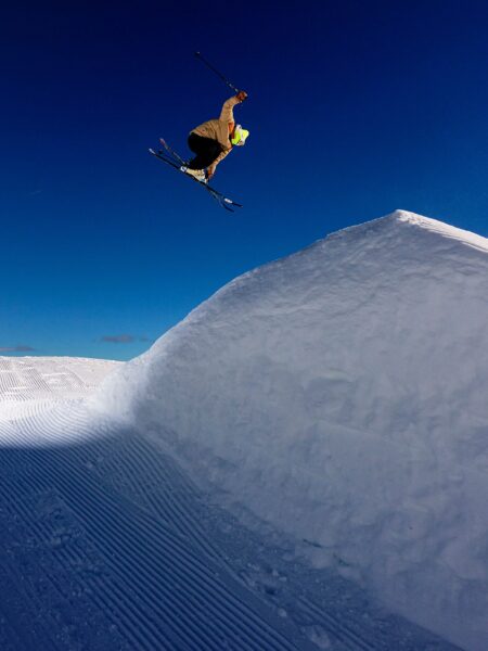 Skier Patrick Ausweger performing a mid-air trick against a clear blue sky, above a large snow jump with groomed tracks on the slope below.