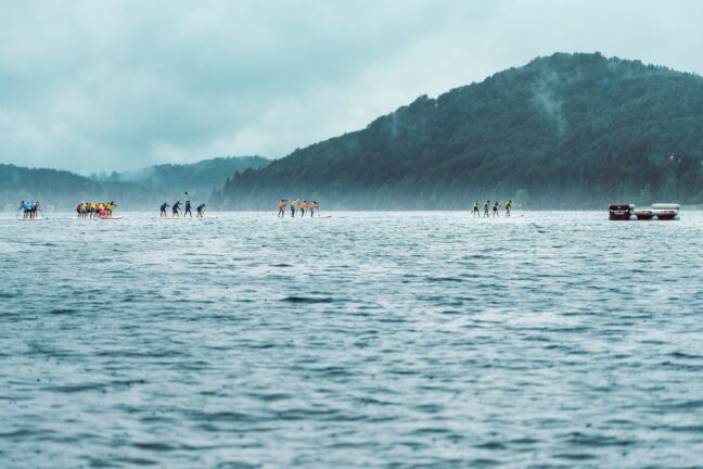 Groups of people participating in a stand-up paddleboarding event on a misty body of water with forested hills in the background.