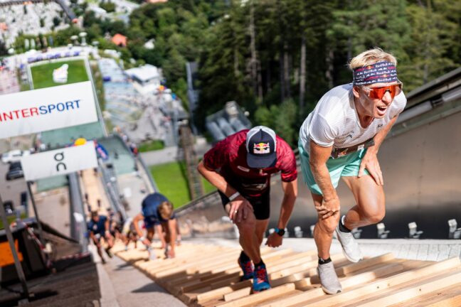 Competitors race up steep wooden planks at the Red Bull 400, with one athlete in the foreground wearing a headband and sunglasses leading the ascent.