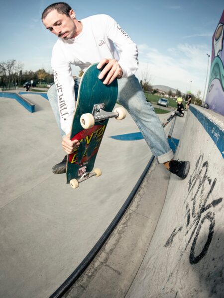 A skateboarder performs a trick at the edge of a skate park ramp, holding the tip of the board with one hand, while dressed in casual attire with a clear sky overhead.