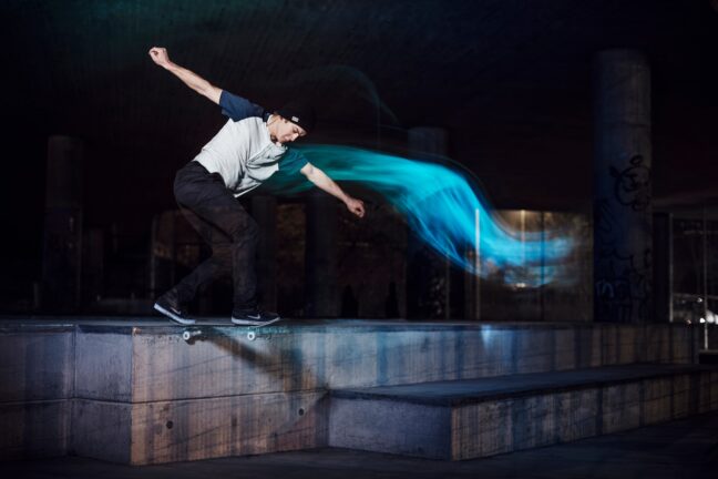 A skateboarder performs a trick on a concrete ledge at night with a blue light trail flowing from his arm, creating a dynamic sense of motion, in an urban skatepark setting.