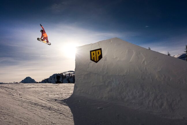 A snowboarder performing a high jump off a snowy ramp against a clear blue sky, with sunlight creating a silhouette effect from behind the ramp.