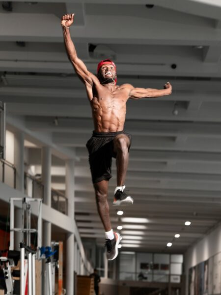 Will Claye in black shorts, a red cap, and running shoes jumping exuberantly with one arm stretched upward inside an indoor gym with weight training equipment in the background.