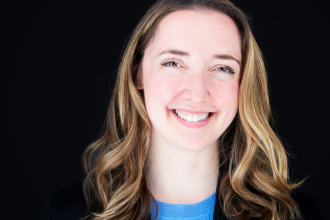 A smiling woman with shoulder-length wavy hair, wearing a blue top and black blazer, against a black background.