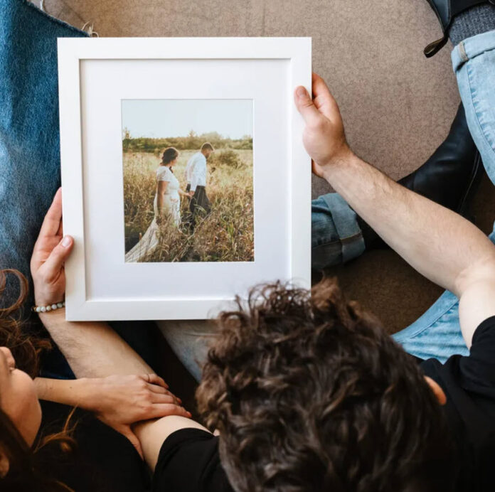 A couple holding hands while sitting on the floor, with one person holding a framed photo of a man and a woman standing in a field printed by Canvaspop.