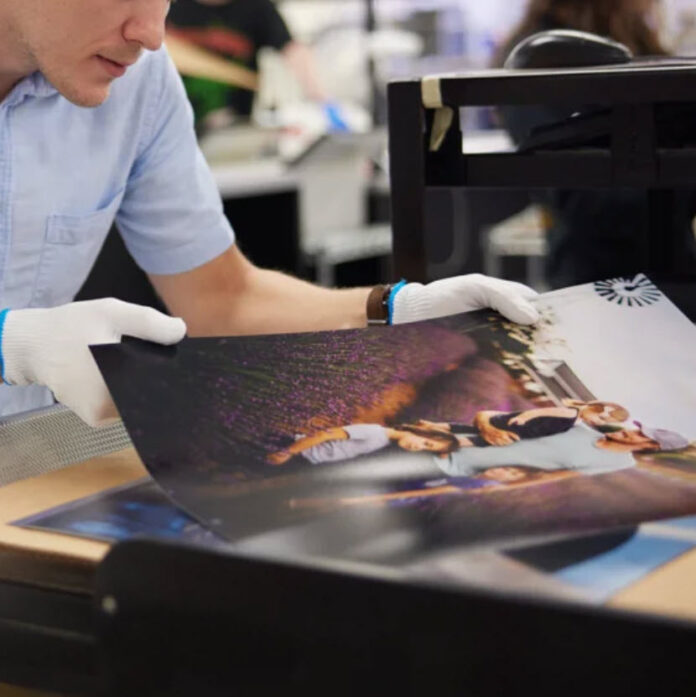 Mpix photo printing staff wearing gloves inspects a large printed photo of two individuals sitting among purple flowers.