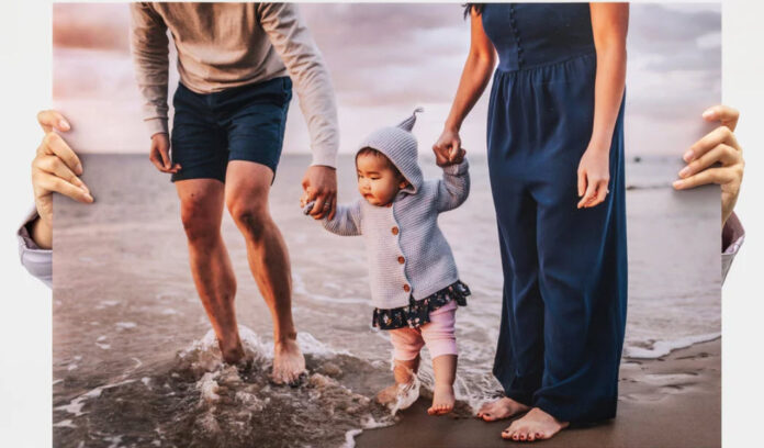 Nations Photo Lab print of a toddler in a gray hooded sweater and pink leggings holding hands with adults on either side while walking on a sandy beach with waves washing over their feet.